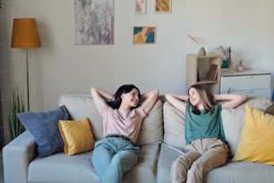 Cheerful and relaxed mother and daughter in casualwear looking at each other while sitting on soft couch and chatting in home environment