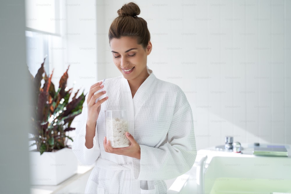 Happy lady in a bathrobe standing near the bathtub and smiling while opening a glass jar of sea salt
