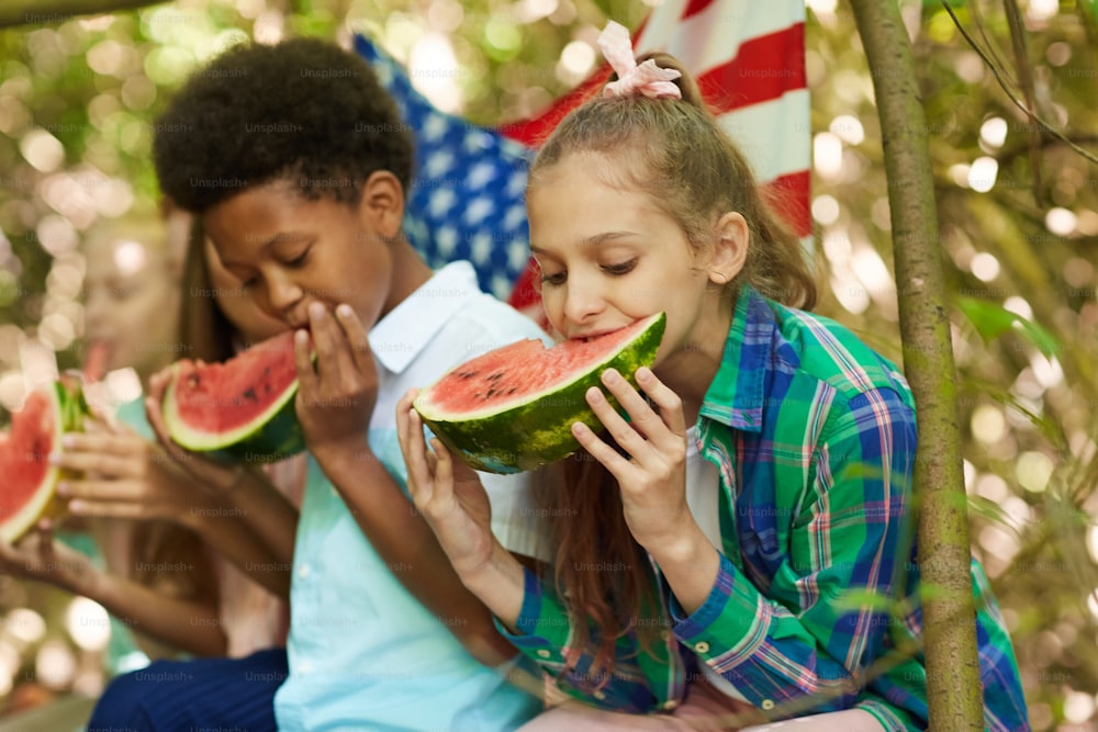 Multi-ethnic group of kids eating watermelon sitting in row while playing outdoors in Summer, focus on cute girl in foreground, copy space