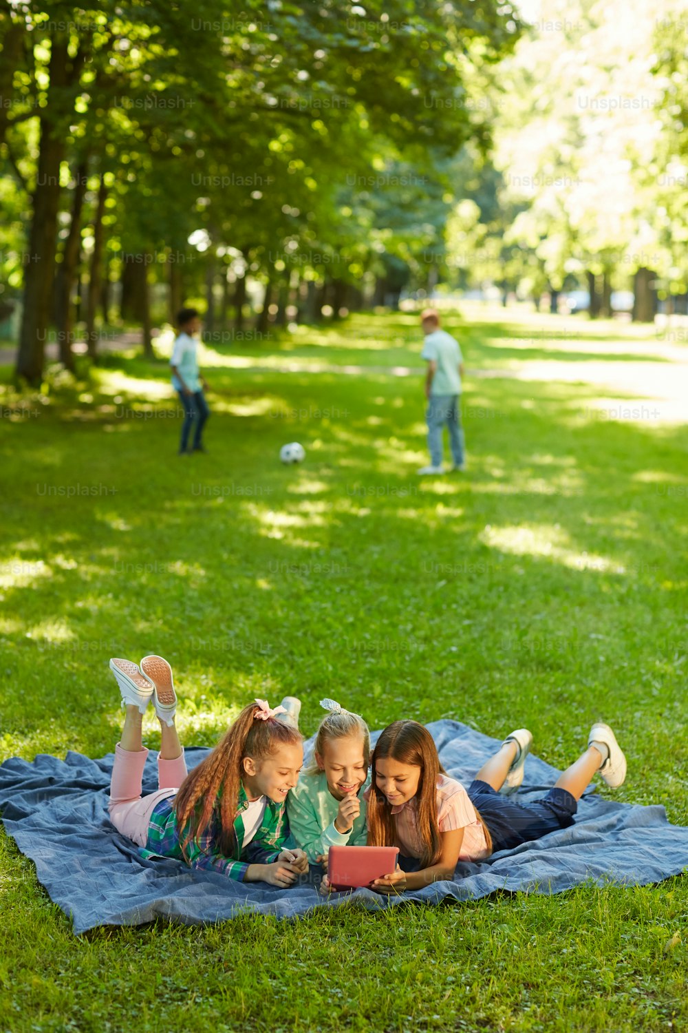 Portrait vertical en pied de trois adolescentes utilisant une tablette numérique allongées sur de l’herbe verte dans un parc à l’extérieur éclairé par la lumière du soleil, espace de copie