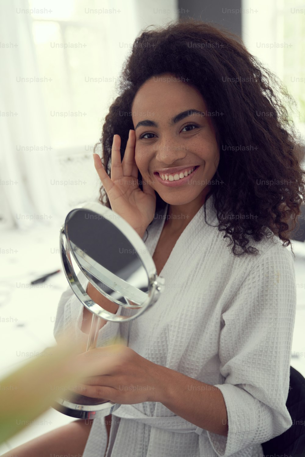 Waist up portrait of cheerful curly female standing in bathrobe and holding hand mirror at home