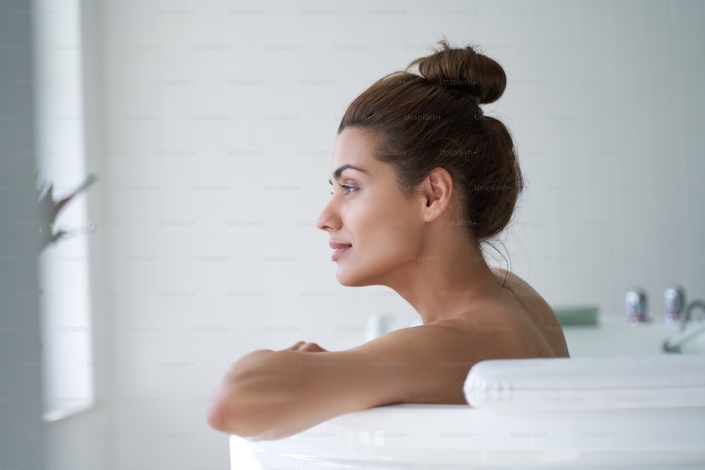 Vista lateral de una mujer joven mirando a lo lejos mientras está sentada en una bañera en el salón de spa
