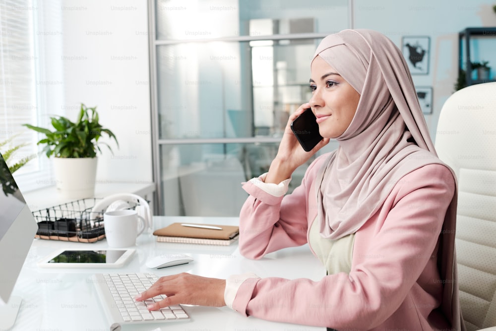 Content attractive young Muslim businesswoman in beautiful hijab communicating by cell phone in office