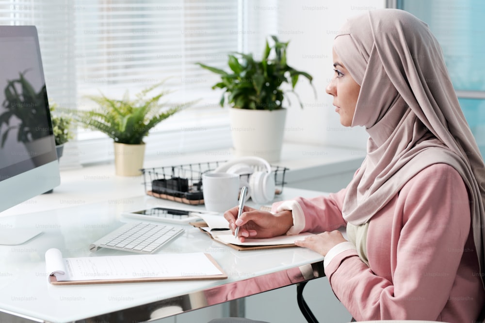 Giovane donna musulmana indaffarata in hijab seduta in un ufficio moderno e prendere appunti mentre lavora con i dati