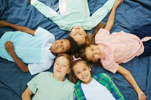 Vista dall'alto al gruppo multietnico di bambini sdraiati in cerchio sulla coperta all'aperto e guardando la macchina fotografica