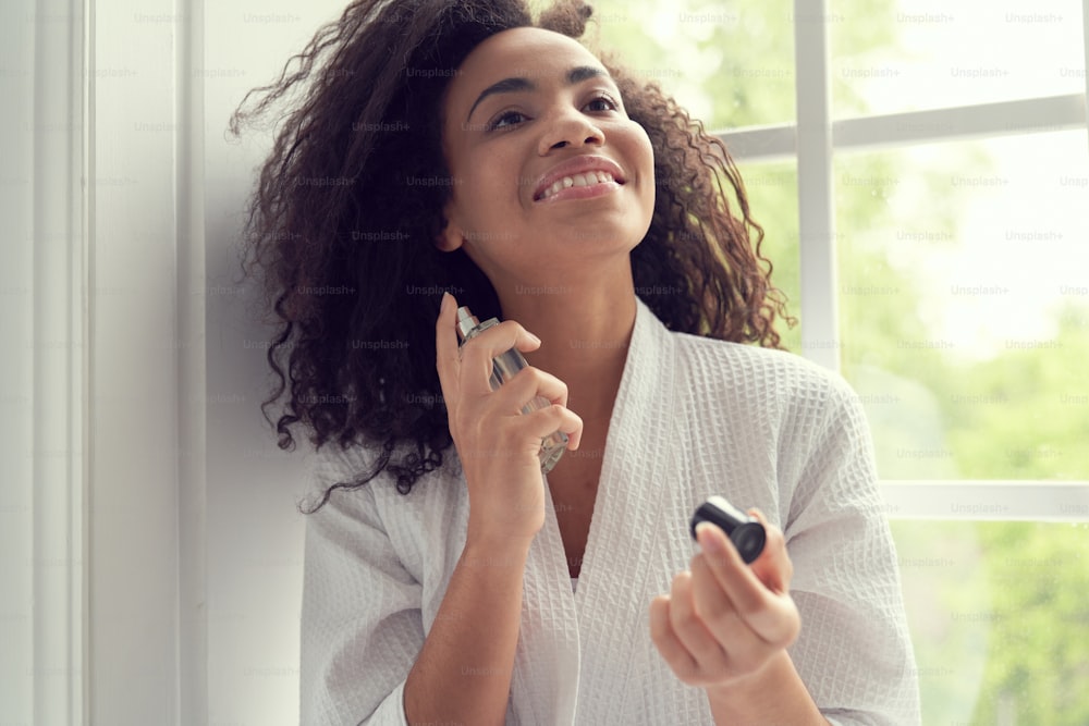 Smiling beautiful curly female is holding bottle of perfume and spraying it on body indoors