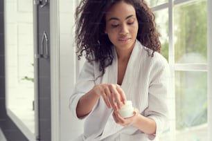Calm beautiful young woman is holding cream for face while looking after skin at home