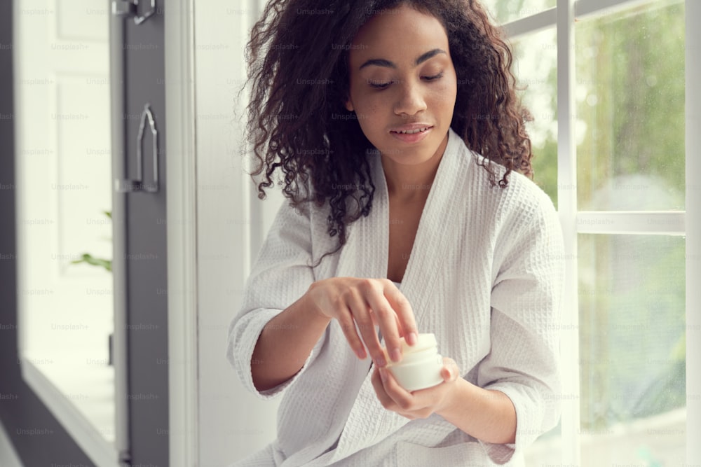 Calm beautiful young woman is holding cream for face while looking after skin at home