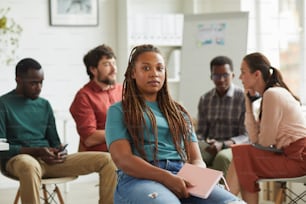 Multi-ethnic group of people sitting in circle while discussing business project in office, focus on African-American woman looking at camera in foreground, copy space