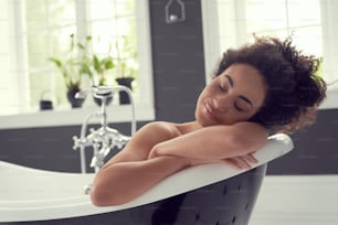 Ruhige lockige Frau schließt die Augen mit Vergnügen, während sie Freizeit genießt und ein Bad nimmt