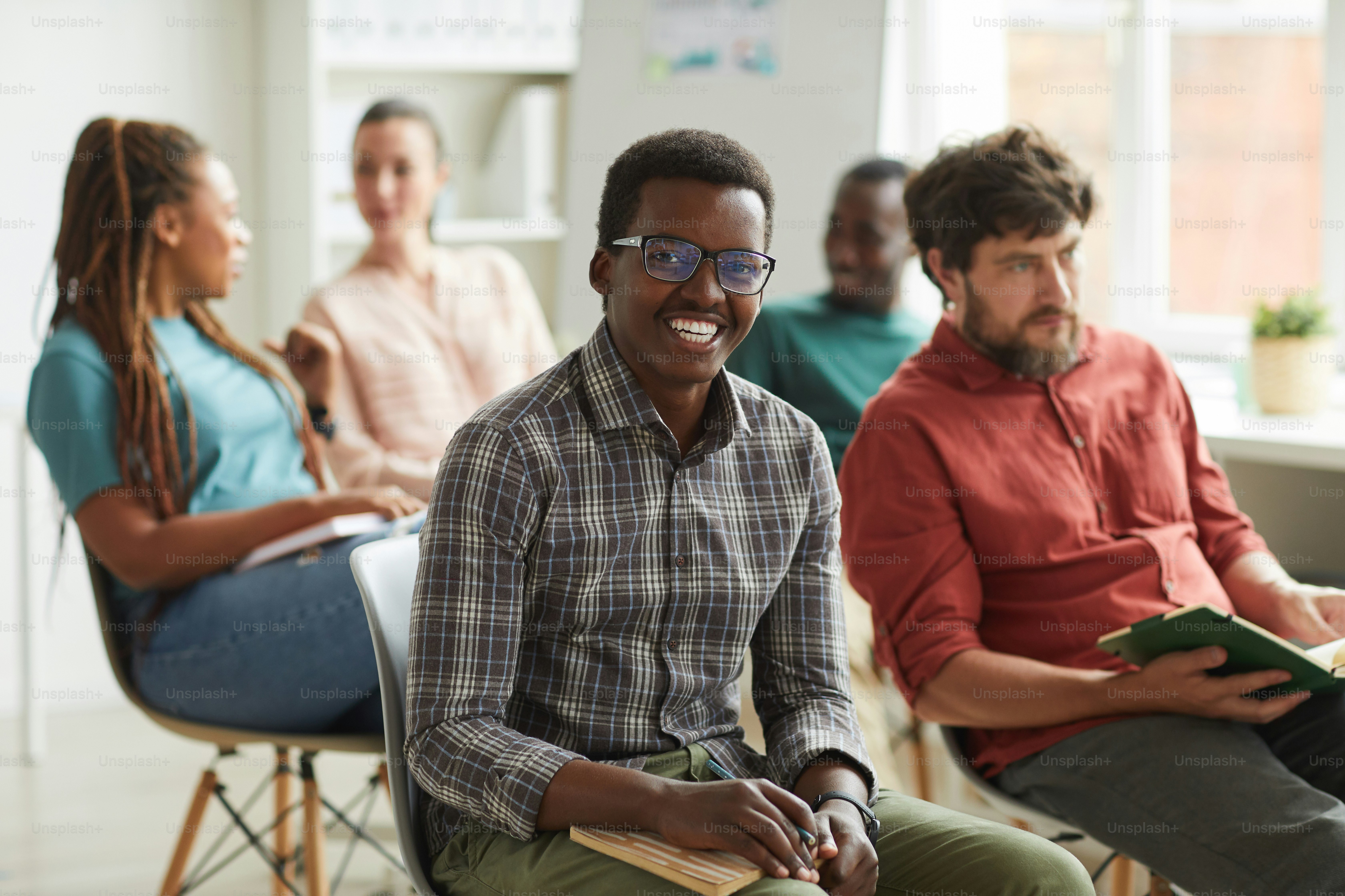 Grupo multiétnico de pessoas sentadas na plateia durante seminário de treinamento ou conferência de negócios no escritório, foco em jovem afro-americano sorrindo para a câmera em primeiro plano, espaço de cópia