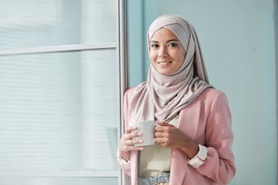Portrait of smiling young Muslim woman in pink jacket standing in office and drinking coffee from mug
