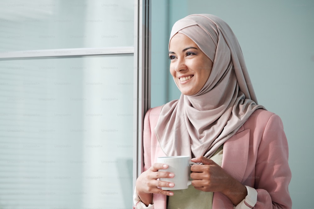 Cheerful attractive young Muslim woman in pink hijab standing at door and holding mug while looking away