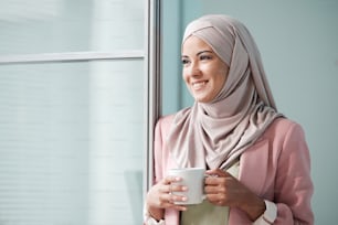 Joyeuse jeune femme musulmane en hijab rose debout à la porte et tenant une tasse tout en détournant le regard