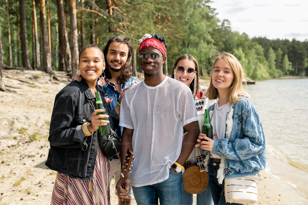 Retrato de jóvenes amigos interraciales alegres de pie con botellas de cerveza en la playa