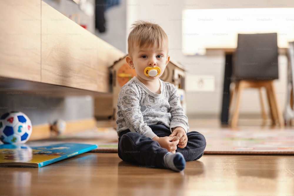 Cute little boy sitting at home with pacifier in his mouth.