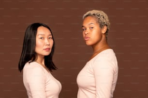 Two serious girls of African and Asian ethnicities in white pullovers standing against brown background in front of camera in isolation