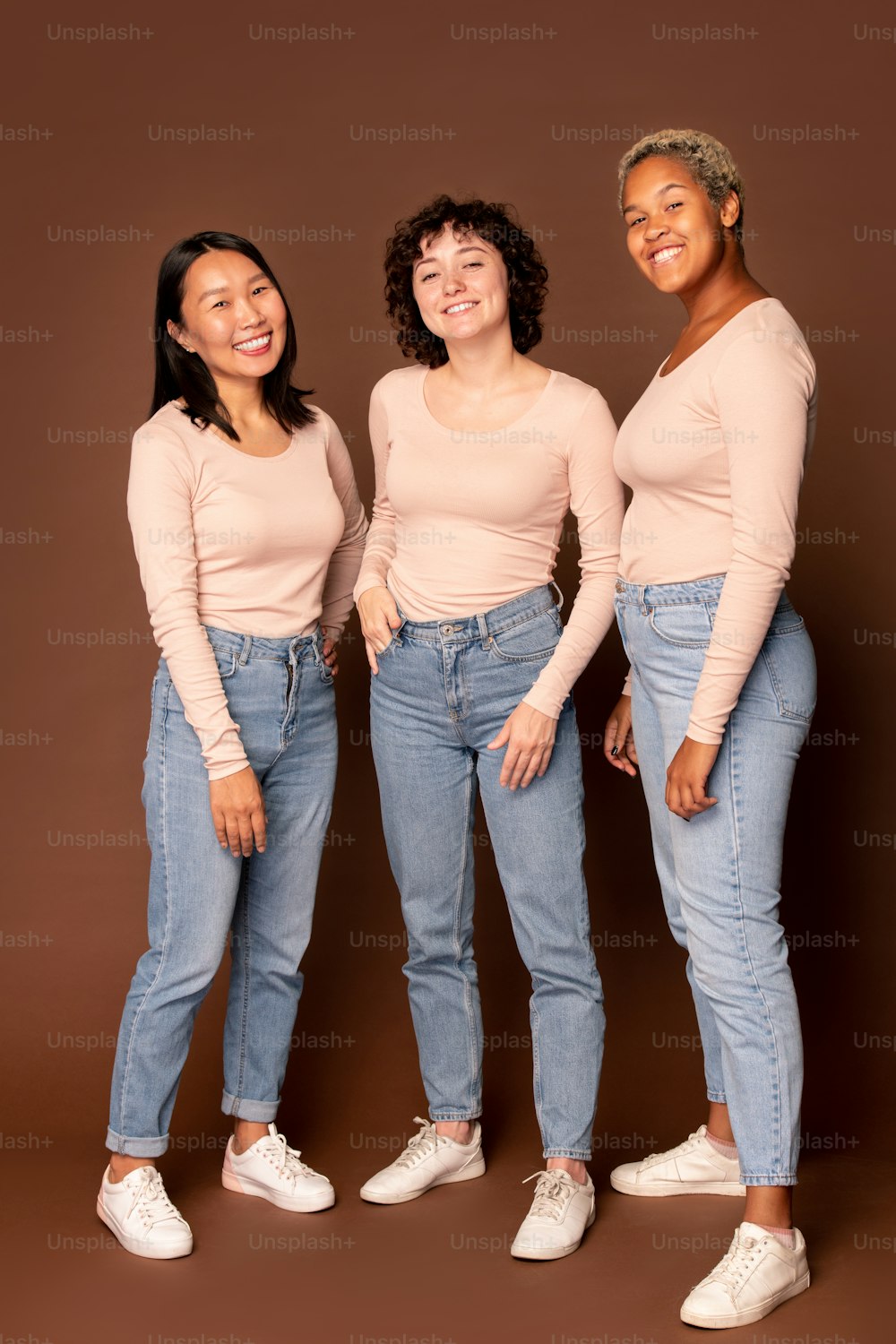 Three young intercultural joyful women in white pullovers and blue jeans standing close to one another and looking at you with toothy smiles