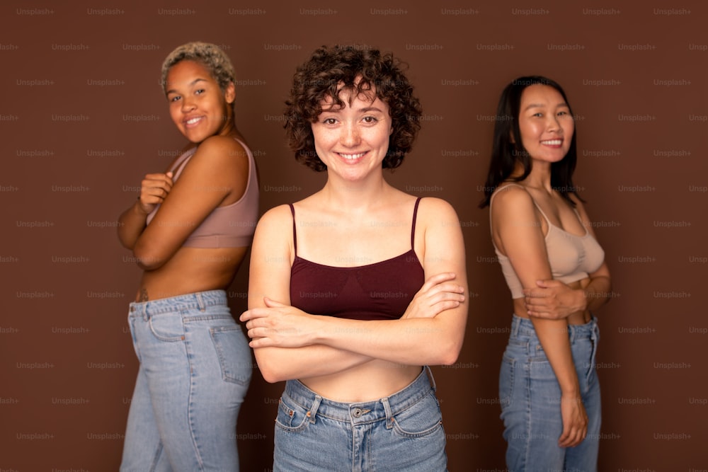 Young beautiful brunette woman in tanktop and blue jeans standing in front of two other intercultural females against brown background
