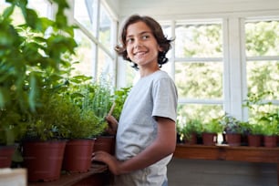 Fröhlicher Junge, der ein Garteninstrument benutzt und lächelt, während er sich zu Hause um Zimmerpflanzen kümmert