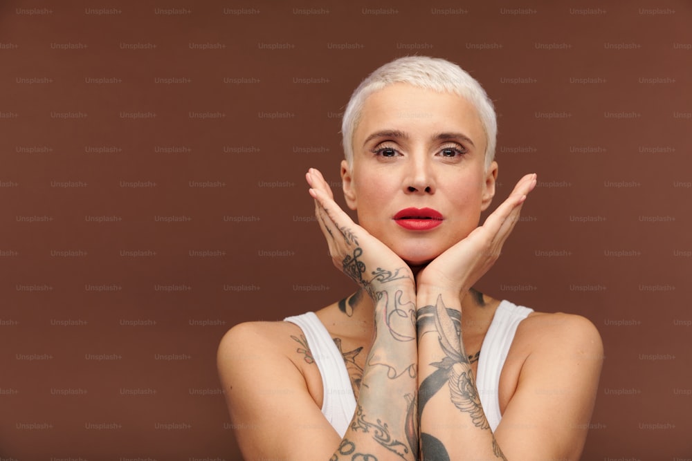 Contemporary young female with short blond hair and tattooes on arms keeping her hands by face while standing in front of camera
