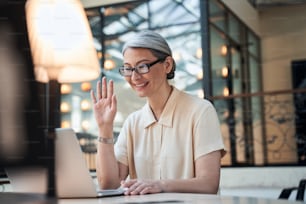 Entusiasta anciana empresaria sentada frente a una computadora portátil agitando la mano mientras tiene una videollamada con sus compañeros de trabajo