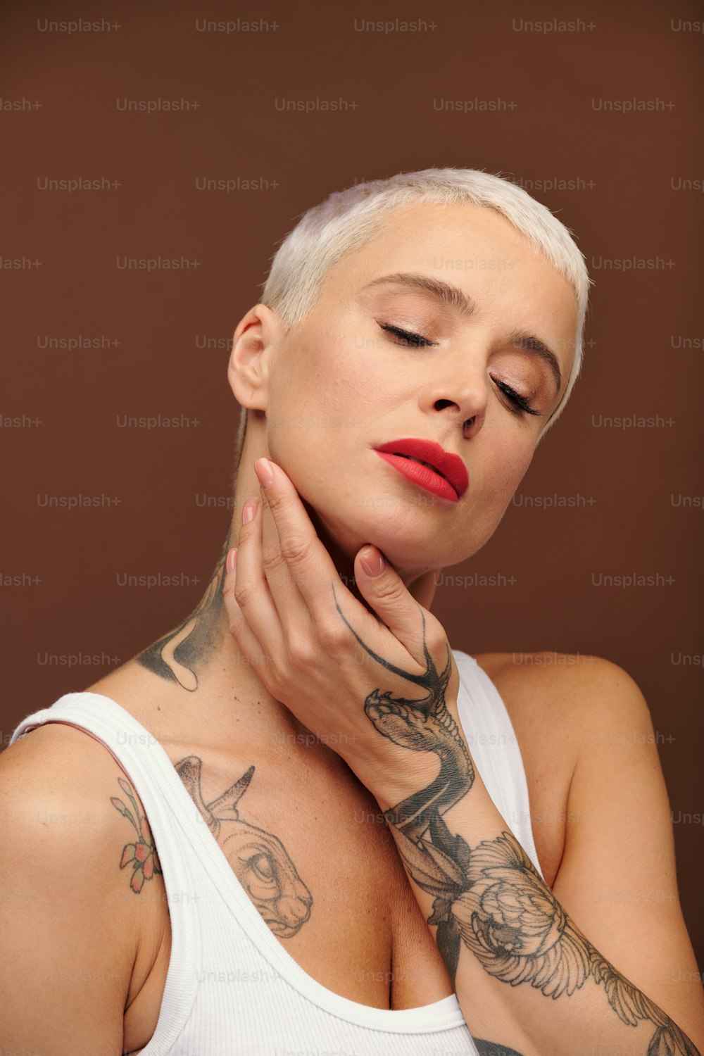 Mature blond female with tattooes on arm touching her face by hand and keeping her eyes closed while standing in front of camera in isolation