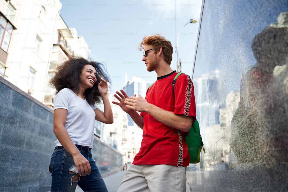 Low angle of cheerful young friends talking while walking in urban street on sunny day