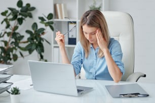 Jeune femme d’affaires fatiguée avec des maux de tête touchant la tempe et se penchant légèrement en avant sur le bureau tout en essayant de se concentrer sur le travail
