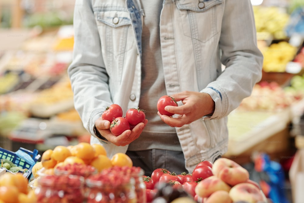 フードカウンターに立ち、オーガニックマーケットで新鮮なトマトを取るジャケットを着た認識できない男性の接写