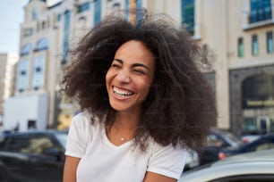 Low angle of attractive curly freckled female laughing while walking in urban street on summer day