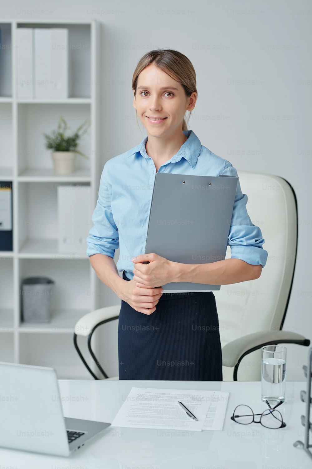 Contemporary young office worker or secretary in formalwear holding clipboard with financial document while standing by workplace