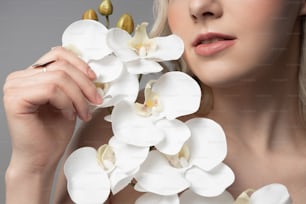 Primer plano de una dama con una piel perfecta tocando pétalos de orquídeas mientras está de pie contra un fondo gris