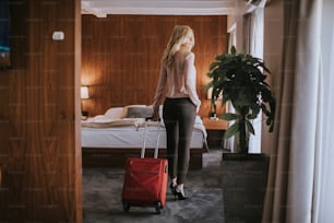 Hübsche junge blonde Geschäftsfrau kommt mit rotem Koffer in ein Hotelzimmer