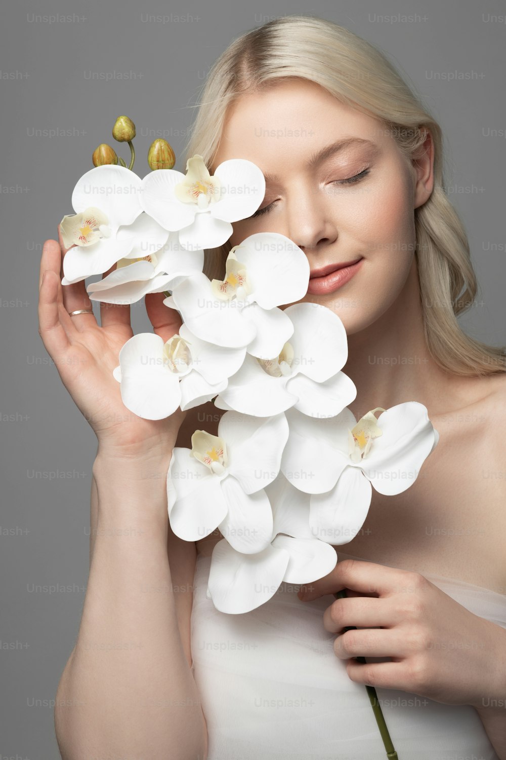 Serene pretty lady with orchid in her hand closing eyes and smiling while standing against gray background