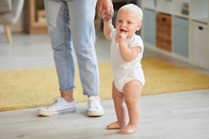집에서 걷는 법을 배우는 아기 아들의 작은 손을 잡고 알아볼 수 없는 여성의 가로 샷