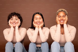 Three young cheerful women of various ethnicities looking at you with toothy smiles while sitting in row with their hands touching face