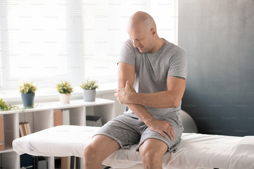 Bald man touching painful elbow and trying to massage it before physical exercise while sitting on medical couch in clinics and waiting for doctor