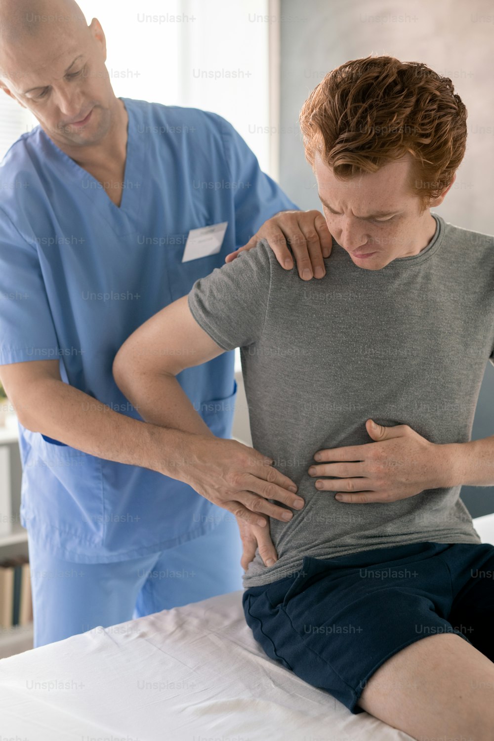 Joven mostrando al médico dónde le duele y tocándose la espalda mientras está sentado en el sofá médico durante el entrenamiento de rehabilitación en clínicas