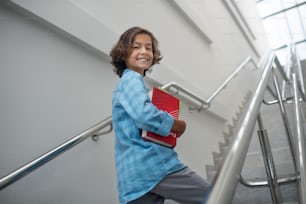 School time. Happy schoolboy holding pile of books, walking upstairs