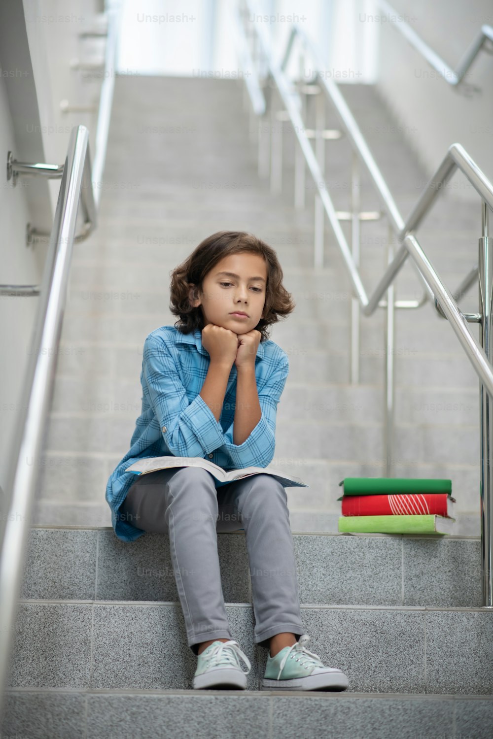 Sadness, mood. Sad boy in light blue rtsbashka and gray trousers sitting on steps of city staircase with book resting his hands on it