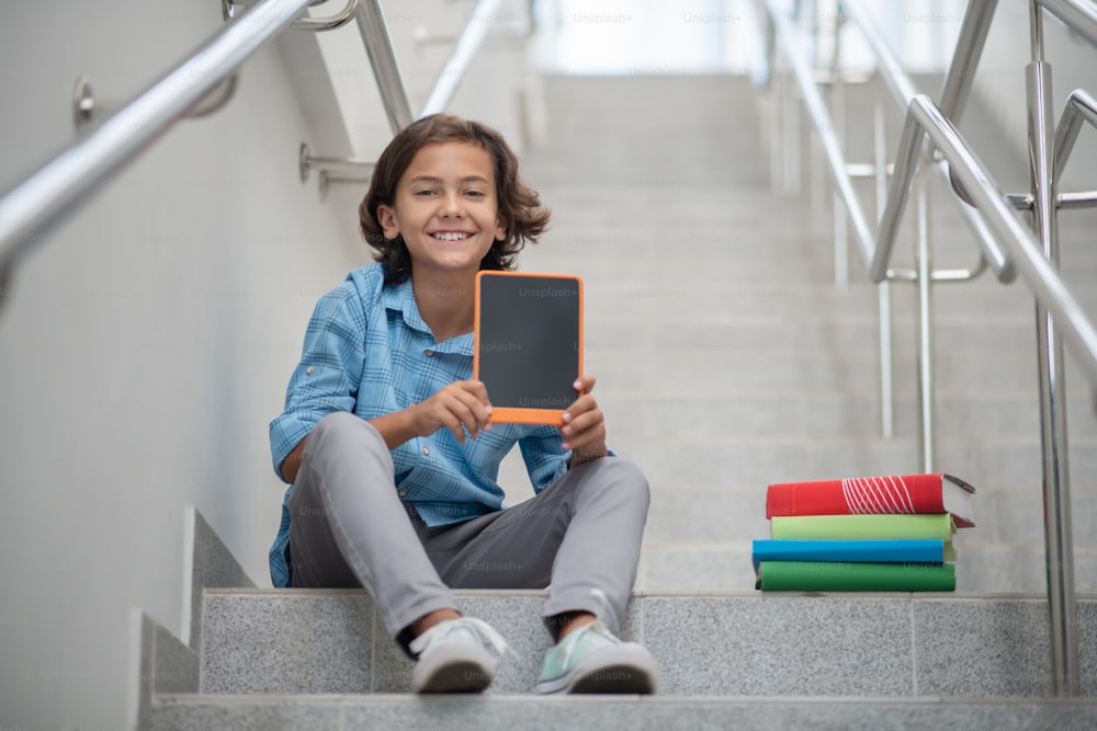 School time. Smiling schoolboy sitting on stairs, showing orange tablet to camera