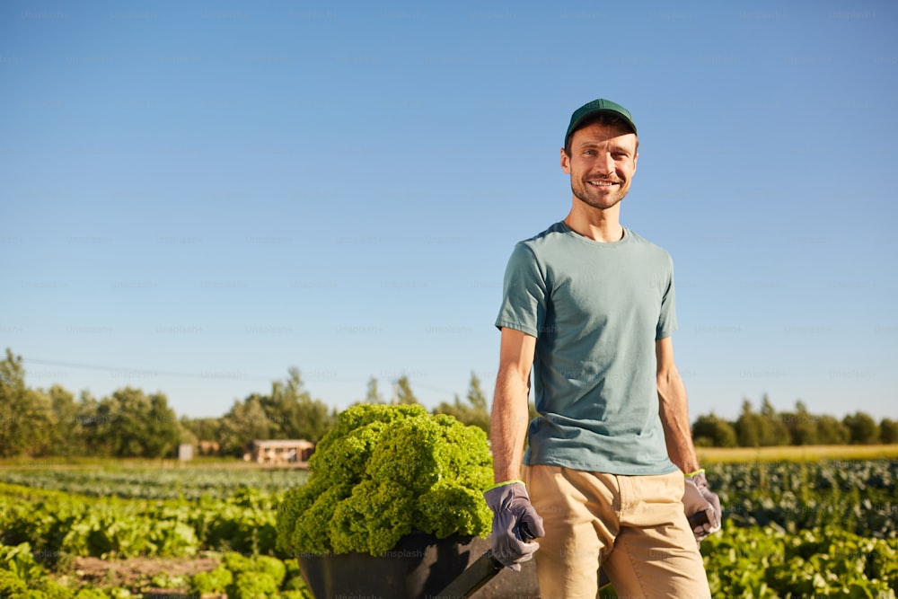Retrato de un trabajador sonriente tirando de un carro cargado con una rica cosecha y mirando a la cámara mientras está parado en una plantación de hortalizas al aire libre a la luz del sol, copie el espacio