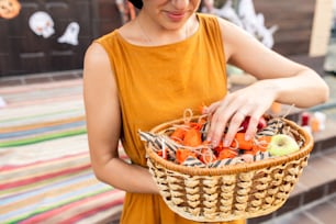 Young elegant woman in yellow dress holding basket with halloween treats wrapped into striped and orange wrappers prepared for children