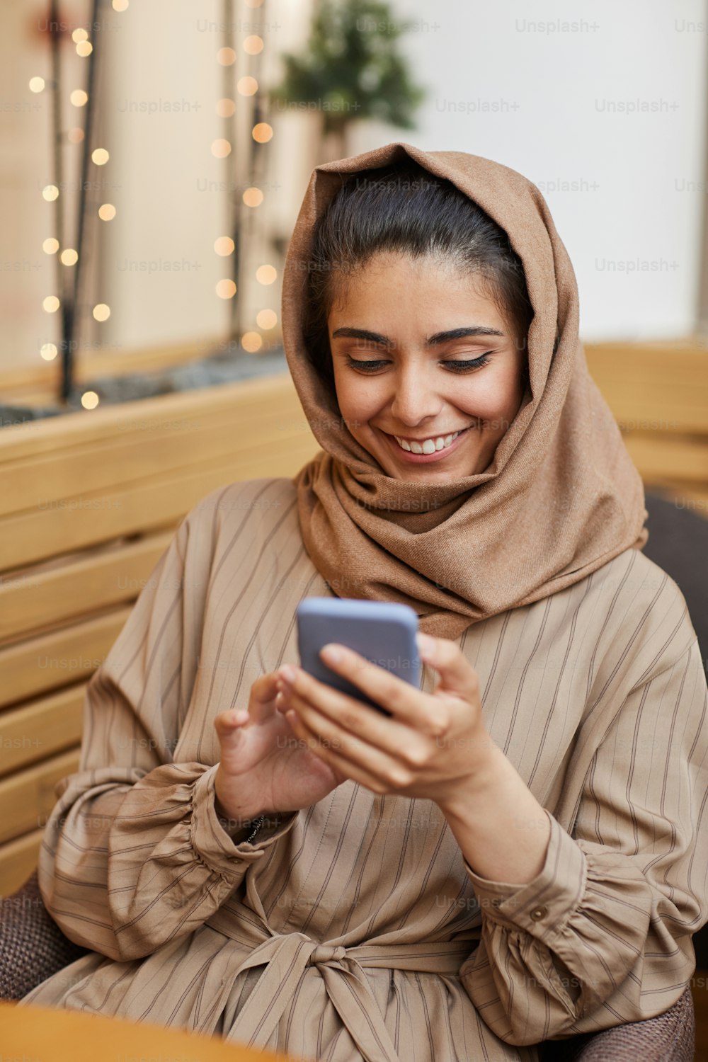 Retrato vertical de una hermosa mujer de Oriente Medio sonriendo felizmente mientras mira la pantalla del teléfono inteligente en un café