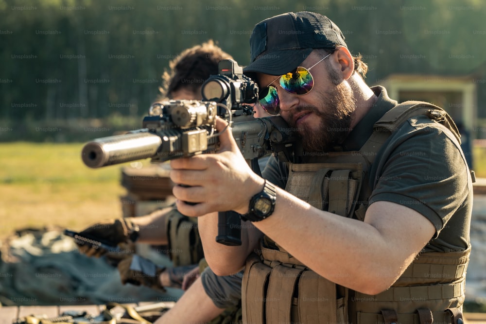 Homem brutal focado em óculos escuros e boné olhando através da mira do rifle enquanto atira no alvo ao ar livre