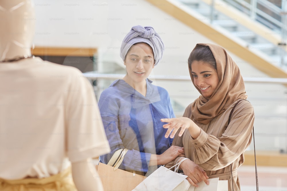 Waist up portrait of two young Middle-Eastern women pointing at showcase display while window shopping in mall, copy space