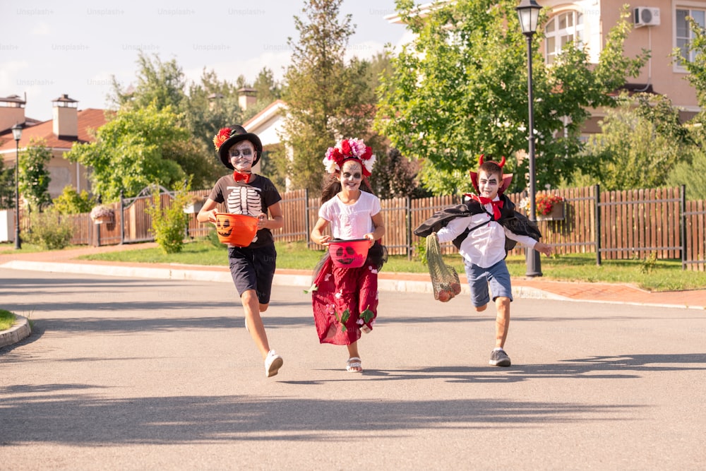Three joyful kids in halloween costumes holding containers with sweet treats while running down wide road on sunny autumn day