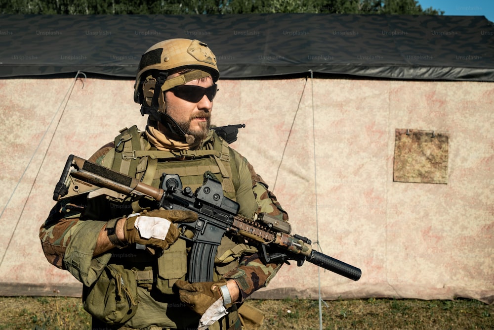 Soldado barbudo confiado en traje de camuflaje y casco sosteniendo un rifle y mirando a lo lejos
