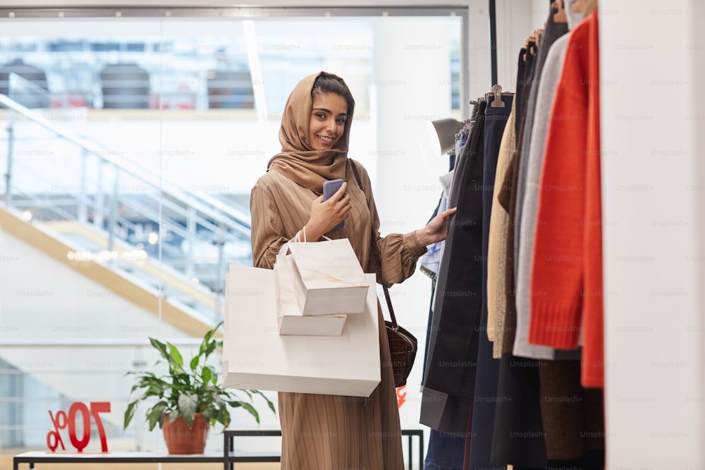 Waist up portrait of elegant Middle-Eastern woman looking at camera while standing by clothing racks and enjoying shopping in mall, copy space